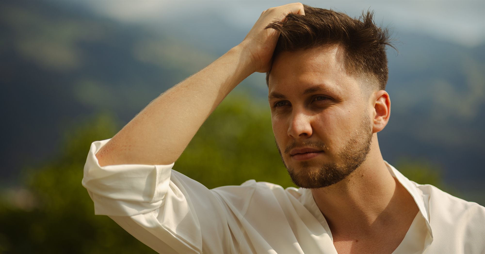 Portrait de Marc en chemise blanche, la main dans les cheveux, devant les montagnes